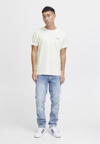 Light yellow cotton t-shirt with small graphic on chest, paired with light blue denim jeans and black-striped white sneakers.