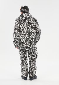 Zwart-wit gemarmerde wintersuit met oversized tekst. Beschikt over een capuchon, grote zakken en waterdicht materiaal. Draagt bij zwarte sneeuwlaarzen.