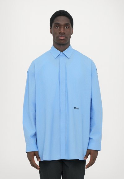 Svetlo modra oversized srajca, mešanica bombaža, z zapiranjem spredaj na gumb, dolgimi rokavi in logotipom na prsih. Gladka tekstura.