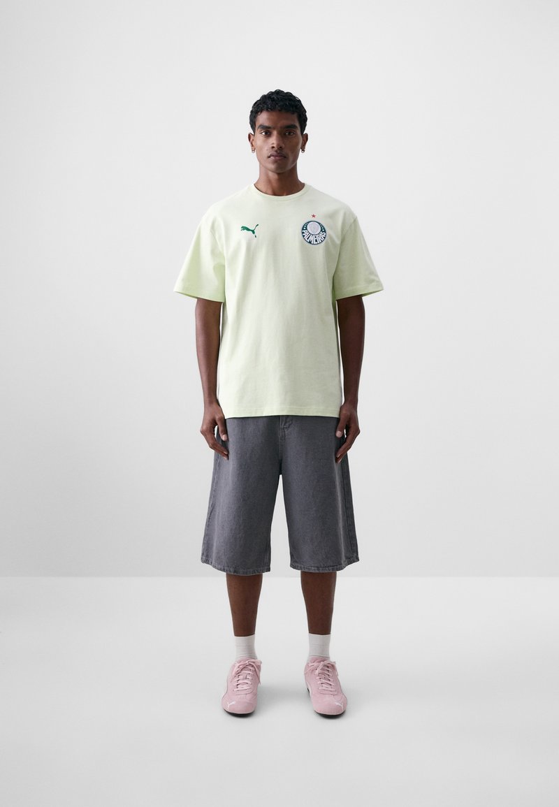Camiseta de algodón verde claro con mangas cortas y logo verde, combinada con pantalones cortos grises y zapatillas deportivas rosas. Corte simple y relajado.