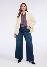 Jeune femme portant un blazer crème, un chemisier à motifs, un jean bleu à larges jambes et des bottes noires à talon pointu, se tenant avec une main dans la poche.