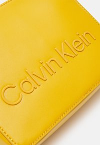 Gul læderpung med præget "Calvin Klein" logo, der har en glat tekstur og kontrasterende syninger langs kanterne.