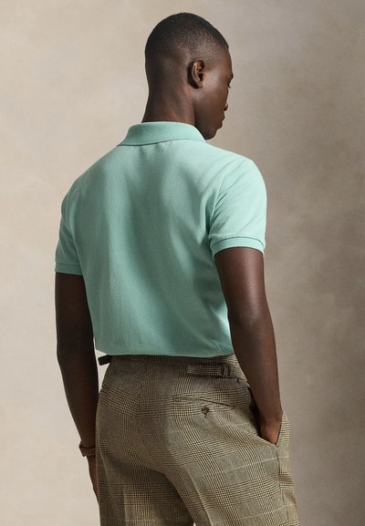 Άνδρας που φαίνεται από πίσω, φοράει ανοιχτό πράσινο κοντομάνικο πόλο πουκάμισο και καρό καφέ παντελόνι με τα χέρια στις τσέπες, με ουδέτερο φόντο.