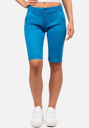 Mujer con pantalones cortos azul brillante hasta la rodilla con bolsillos delanteros y zapatillas blancas, de pie con la mano en la cadera.