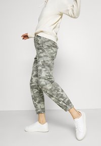 Spodnie cargo w wzór kamuflażu, wykonane z lekkiego materiału, z elastycznymi mankietami, kieszeniami bocznymi i detalami z guziczkami. Noszone z białymi trampkami.