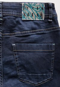 Los jeans de mezclilla azules presentan una etiqueta plateada con un diseño gráfico unida a la cintura, destacando la costura y un detalle del bolsillo trasero.