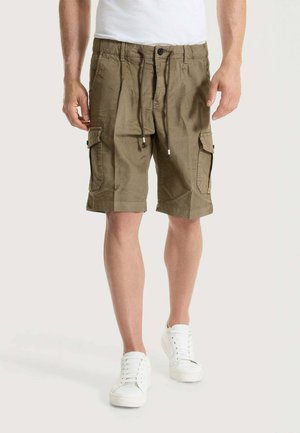 Uomo che indossa pantaloncini cargo verde oliva con tasche laterali, scarpe da ginnastica bianche e una t-shirt bianca, in piedi davanti a uno sfondo chiaro e semplice.