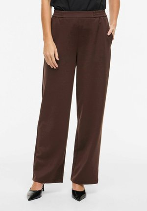 Kobieta w luźnych, ciemnobrązowych spodniach, czarnych szpiczastych szpilkach, stojąca z jedną ręką w kieszeni na białym tle.