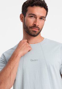 Světle modré tričko s krátkým rukávem, vyšívaným logem "Ombre" a viditelnými švy. Měkká tkanina a uvolněný střih.