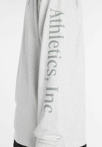 Maglietta a maniche lunghe grigia con la scritta "Athletics, Inc." in un design verticale sulla manica sinistra. Realizzata in tessuto leggero con una texture liscia.