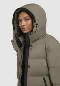 Dik olijfgroene jas met grote capuchon, zwarte accenten, rits- en drukknoopsluiting, en een gestructureerd, gewatteerd stofontwerp.