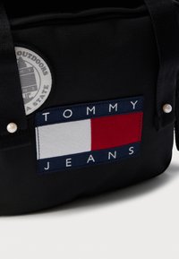 Geantă din material textil negru, cu un petic circular cu logo și o etichetă dreptunghiulară TOMMY JEANS în culorile albastru, alb și roșu. Accentuate cu ținte asemănătoare cu perlele.