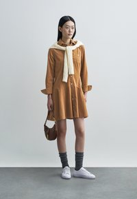 Καφέ φόρεμα από κορδόνι με κουμπιά μπροστά και δύο πλαϊνές τσέπες, σε συνδυασμό με λευκά αθλητικά παπούτσια και γκρι πλεκτές κάλτσες, και κρεμ πουλόβερ δεμένο στον λαιμό.