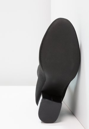 Semelle de botte noire à talon avec un talon texturé, positionnée à la verticale contre un mur et un sol blancs en angle.