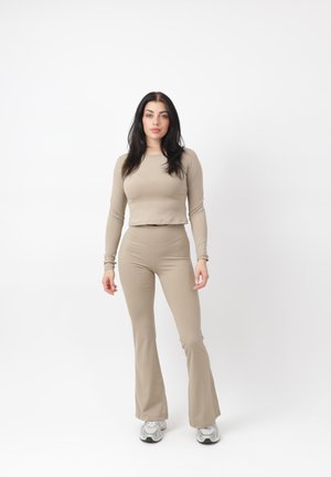 Kvinde med langt mørkt hår stående iført beige langærmet crop top, matchende tætsiddende bukser og hvide sneakers på hvid baggrund.
