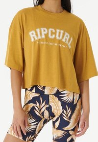 Musztardowa krótka koszulka z białym nadrukiem "RIPCURL". Stylizowana z granatowymi szortami rowerowymi w kwiatowy wzór z jasnymi liśćmi tropikalnymi.