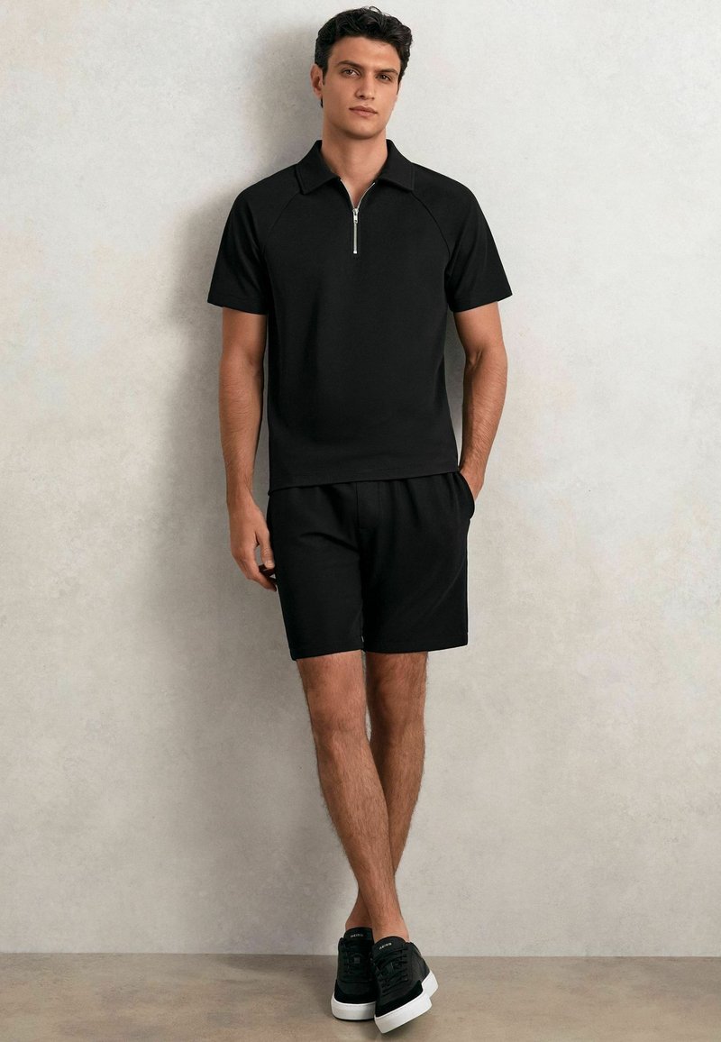 Schwarzes kurzärmeliges Polohemd mit Reißverschlusskragen, kombiniert mit schwarzen Shorts. Beide haben eine glatte Textur und einen lässigen Schnitt. Schwarze Sneakers mit weißen Sohlen.