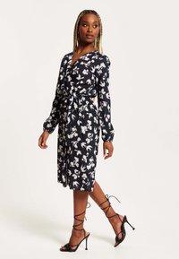 Black dress featuring white floral print, long sleeves, v-neck, and a belted waist. Paired with strappy black high-heeled sandals.