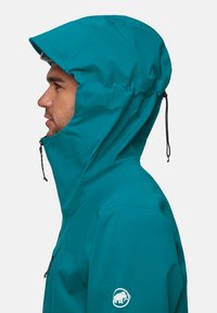 Tee-farbene wasserdichte Jacke mit hohem Kragen und verstellbarem Kapuzen. Verfügt über einen Reißverschluss und ein Logo am linken Ärmel. Glatte, matte Oberfläche.