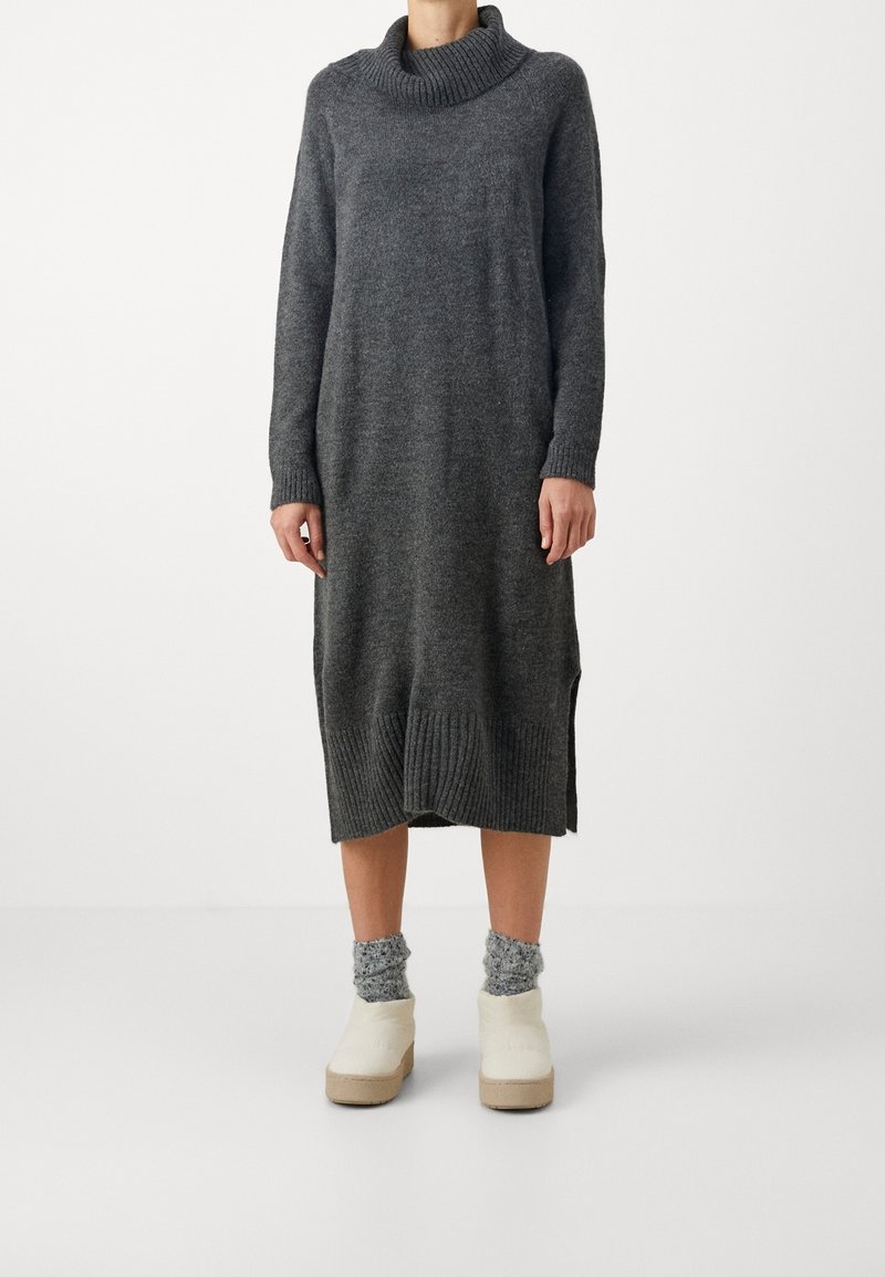 Personne portant une longue robe-pull tricotée gris foncé avec un col roulé roulé, des chaussettes grises et des chaussures compensées blanches slip-on sur fond blanc.