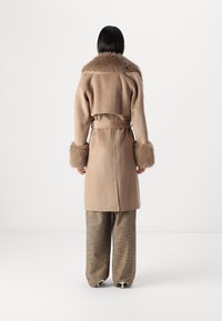 Ανοιχτό καφέ παλτό από μαλλί με γούνινο γιακά και μανσέτες, που έχει ζωνάκι στη μέση. Η πλάτη έχει ίσια κοπή και λεπτομέρειες αεραγωγών. Συνδυάζεται με υφασμάτινο παντελόνι.