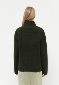 Giacca in pile verde scuro con colletto alto, superficie testurizzata e maniche lunghe, indossata sopra pantaloni chiari e larghi.