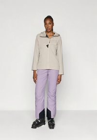 Lichtbeige jas met capuchon en ritssluiting aan de voorkant, gecombineerd met lavendelkleurige broek met marineblauwe accenten en zwarte skischoenen. Eenvoudig, functioneel ontwerp.