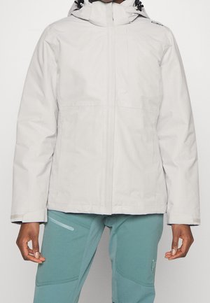 Osoba ubrana w jasnoszarą kurtkę z kapturem i zamkiem błyskawicznym z przodu oraz turkusowe spodnie outdoorowe, stojąca na jednolitym jasnym tle.