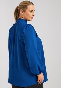 Blusa blu con maniche lunghe a sbuffo e colletto alto. Presenta una apertura a occhiello sul retro e un tessuto texturizzato. Vestibilità sartoriale.