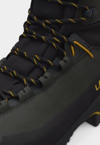 Κοντινό πλάνο ενός μαύρου και σκούρου γκρι παπουτσιού πεζοπορίας με μαύρα κορδόνια με κίτρινα στοιχεία, μαξιλαρωμένη γλώσσα και ανθεκτική υφή στη σόλα.