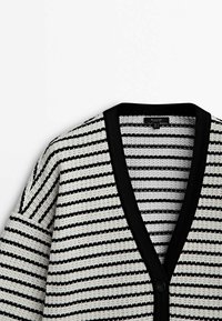 Cardigan tricoté à rayures noires et blanches avec un profond col en V et une bordure noire le long de l'encolure et de la zone des boutons.