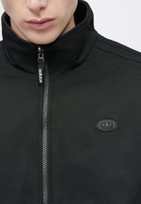 Μαύρο μπουφάν με φερμουάρ και ψηλό γιακά, με υφασμάτινη υφή και ανάγλυφο λογότυπο στο αριστερό στήθος. Μινιμαλιστικός σχεδιασμός.