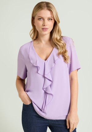 Blonde vrouw met golvend haar, gekleed in een lichtpaars blouse met ruches en een V-hals, en donkerblauwe jeans, staand met één hand in haar zak.