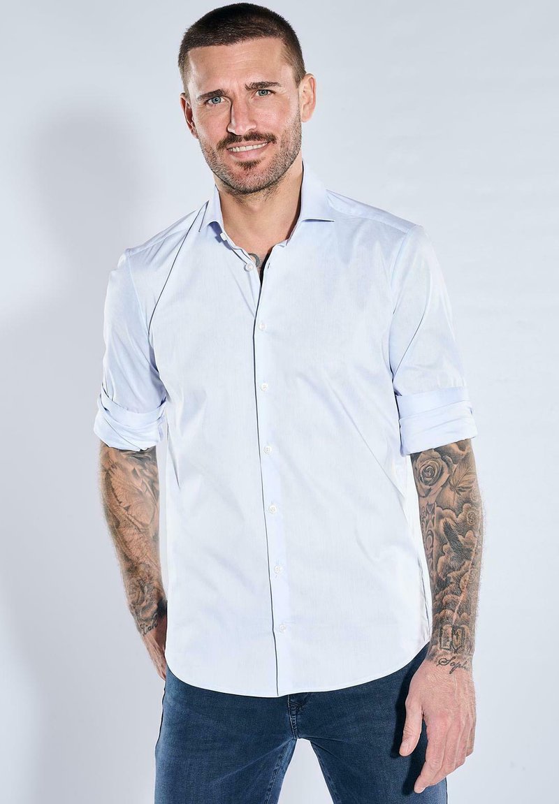 Smiling man with short hair wearing a rolled-up white shirt and dark jeans, showing full-sleeve floral and script tattoos on both arms.