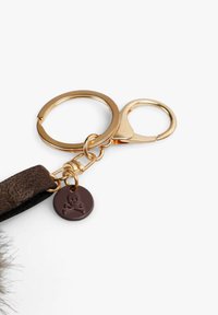 Llave de cuero marrón con un anillo de metal dorado, que presenta un mosquetón dorado y un charm de color marrón oscuro con un diseño de calavera y huesos cruzados.