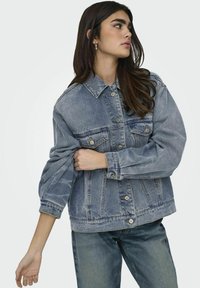 Giacca in denim azzurro chiaro con colletto, vestibilità oversize, tasche frontali e chiusure con bottoni argentati. Polsini a costine e texture leggermente consumata.