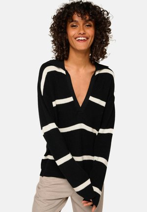 Femme souriante avec des cheveux bouclés portant un pull noir à rayures blanches et un pantalon beige, se tenant devant un fond uni.