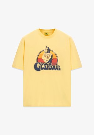 Geel T-shirt met korte mouwen met een afbeelding van een vrouw die een Guess-logo vasthoudt met de tekst "hysteric GLAMOUR" onder een retro zonsondergangcirkel.