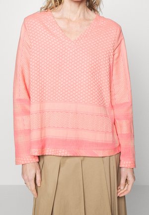 Vrouw die een koraalroze, getextureerde V-hals trui met lange mouwen en een beige geplooide rok draagt, staand tegen een lichte achtergrond.