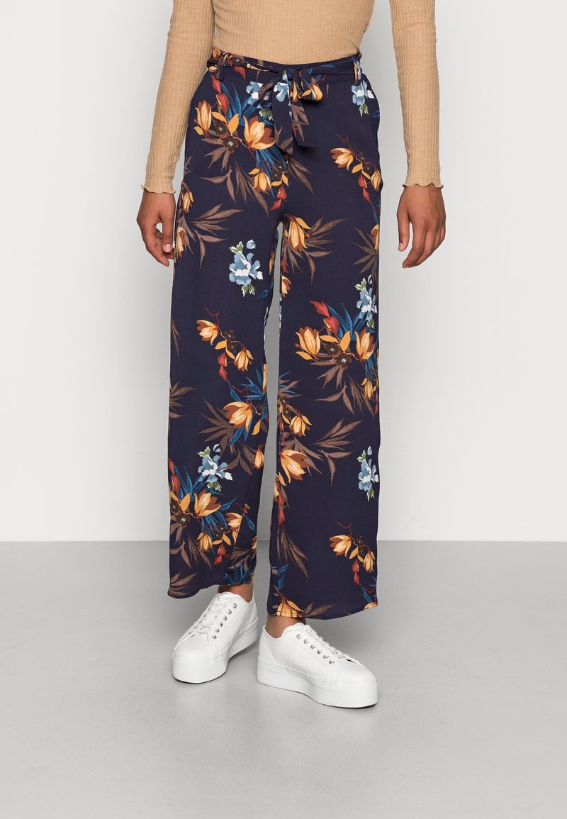 Pantalon large bleu marine avec imprimé floral orange et bleu, taille nouée, porté avec un haut beige à manches longues côtelé et des baskets blanches à plateforme.