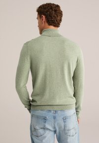 Grüner Pullover mit langen Ärmeln und hohem Kragen. Hergestellt aus weichem, strukturiertem Stoff. Kombiniert mit hellblauen Jeans aus Denim mit Taschen.