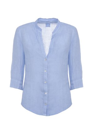 Donna che indossa una camicetta azzurro chiaro con bottoni e pantaloni ampi abbinati, tiene una borsa tote beige intrecciata, in piedi su sfondo bianco.