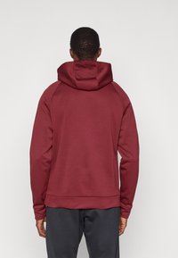 Vyras, vilkintis bordo spalvos džemperį su gobtuvu ir tamsiai pilkas kelnes, parodytas iš nugaros ant vienspalvės šviesios fono.