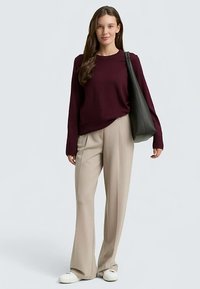 Brązowy sweter z długimi rękawami i okrągłym dekoltem, zestawiony z beżowymi spodniami o szerokich nogawkach. Modelka trzyma czarną torbę typu tote, na stopach białe sneakersy.