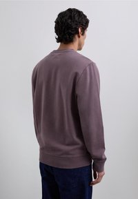Sudadera de algodón morada con cuello redondo, puños y dobladillo acanalados, que presenta un diseño simple y una textura suave, combinada con pantalones de mezclilla oscuros.