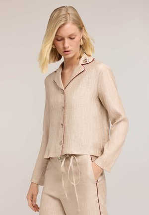 Blonde Frau trägt beige strukturierte, geknöpfte Bluse mit Kragen und passende Kordelhose, Hand in der Tasche, vor einfarbigem Hintergrund.
