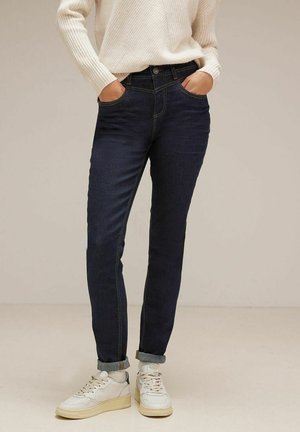 Mørkeblå jeans lavet af denim, med slim fit, fem-lomme design og subtil gul søm. Sammen med hvide sneakers.