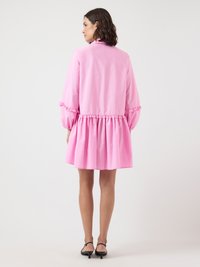 Kvinna vänd bort från kameran, klädd i en rosa långärmad klänning med volangdetaljer och svarta kitten heel-sandaler, framför en enfärgad bakgrund.