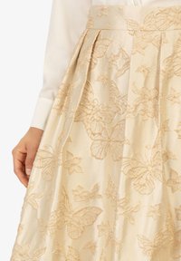 Jupe beige avec broderie florale et de papillons. Présente des plis et une texture lisse et brillante. Association présentée avec un blouse blanche.