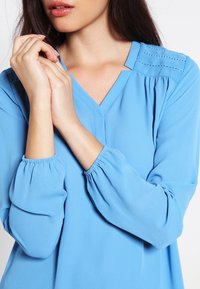 Femme portant un chemisier bleu clair à manches longues avec des poignets froncés et des détails cousus près des épaules, les mains doucement jointes près du visage.